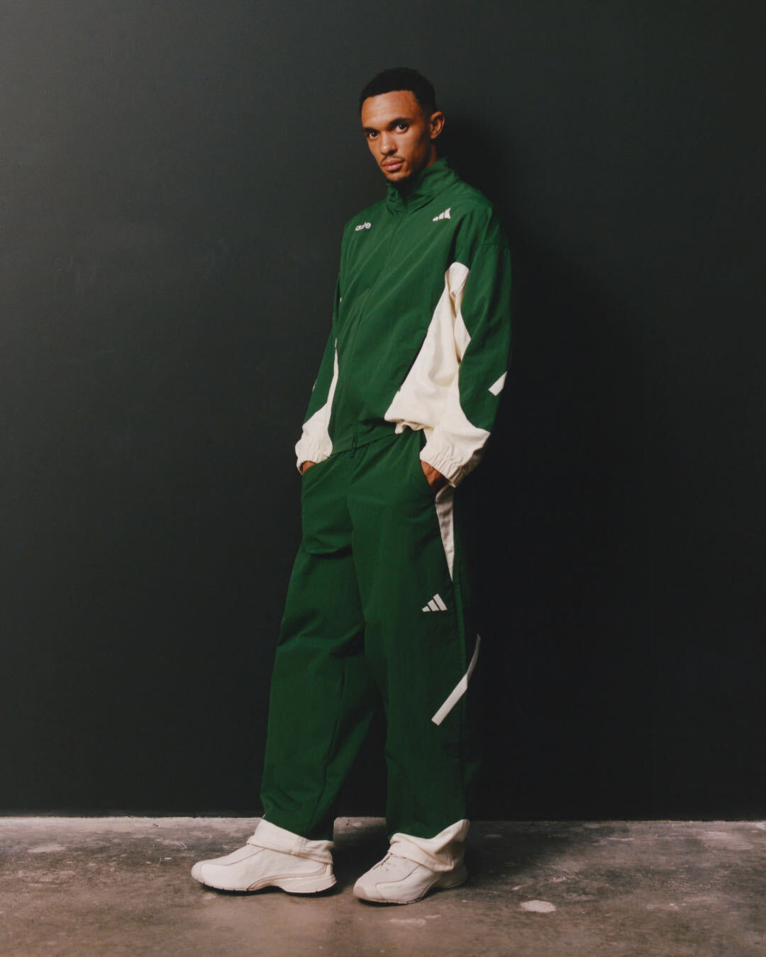 Un homme portant un survêtement Adidas x Arte vert et blanc, composé d'une veste à fermeture éclair et d'un pantalon assorti, assorti à des baskets blanches, debout devant un mur gris foncé.