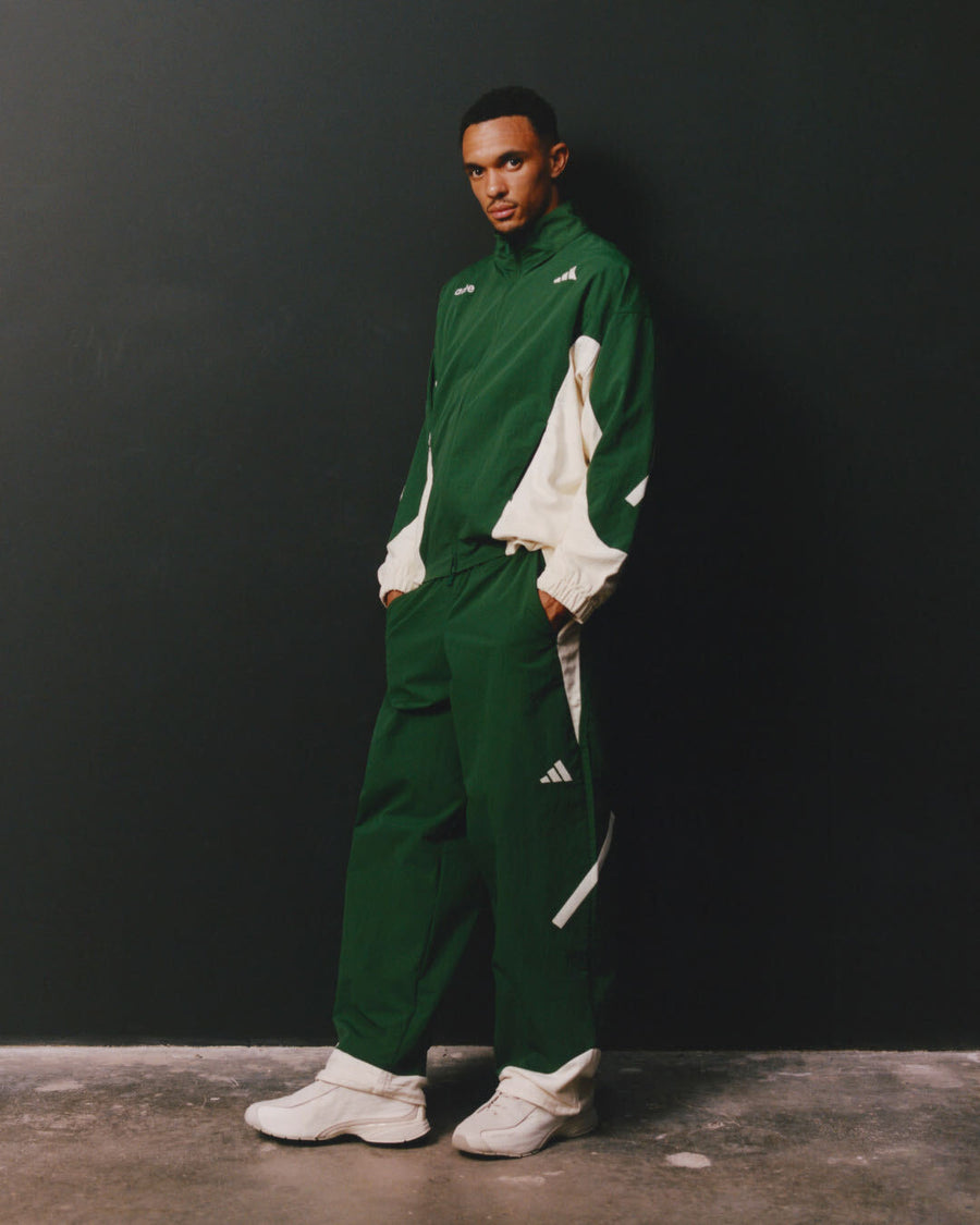 Un homme portant un survêtement Adidas x Arte vert et blanc, composé d'une veste à fermeture éclair et d'un pantalon assorti, assorti à des baskets blanches, debout devant un mur gris foncé.